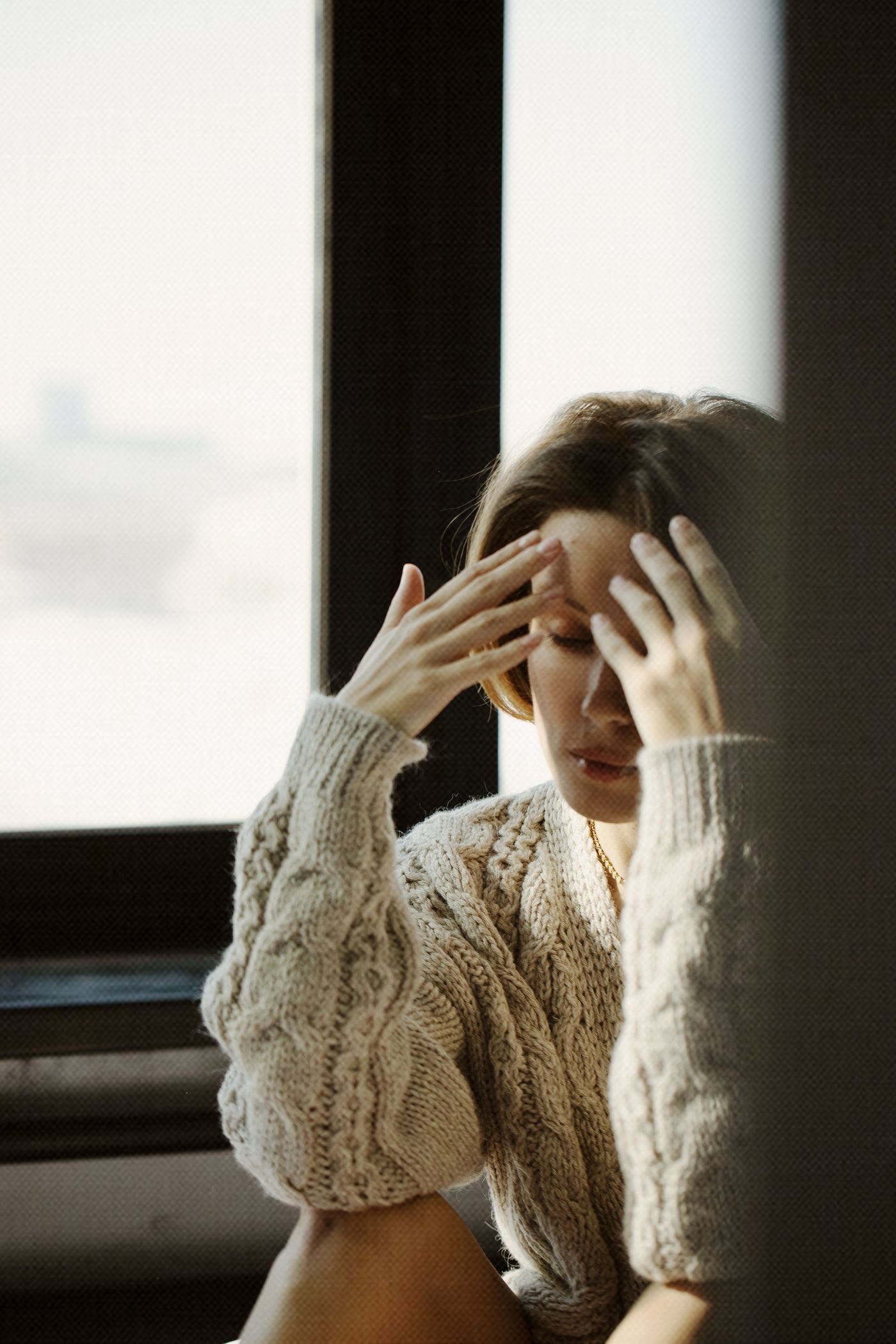 Woman sitting by a window with her hands on her face, lost in thought